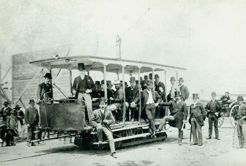 Black and white photo of men in top hats posed around a tram in 1890.