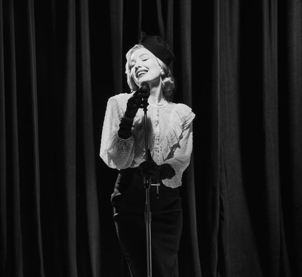 Black and white photo of woman dressed in hat and gloves singing into a microphone, stage curtain in background.