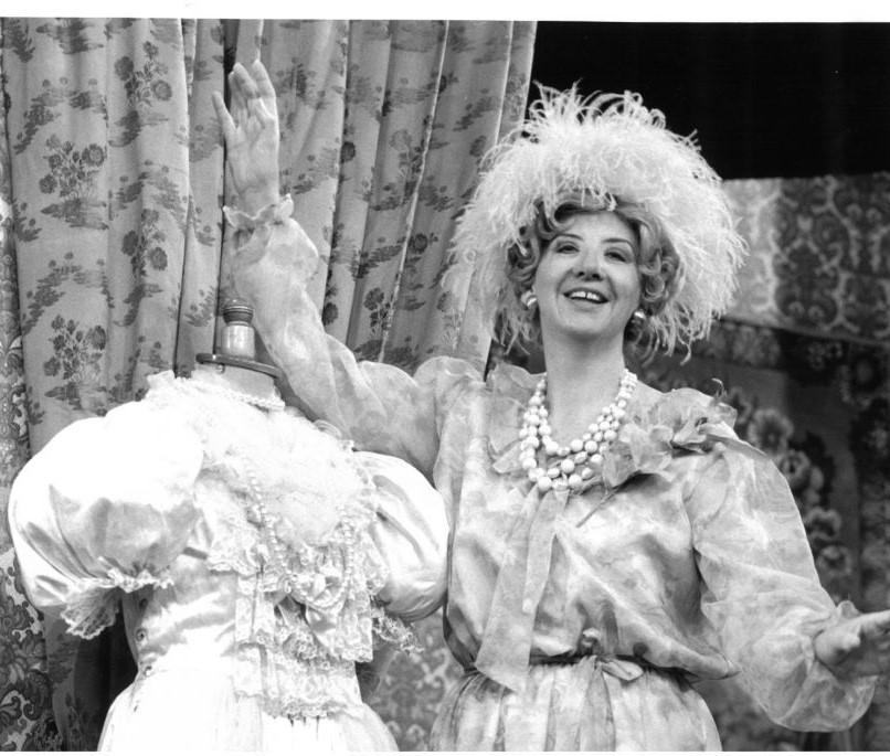 Black and white photo of actress on stage in a feather hat and pearls next to a costume mannequin