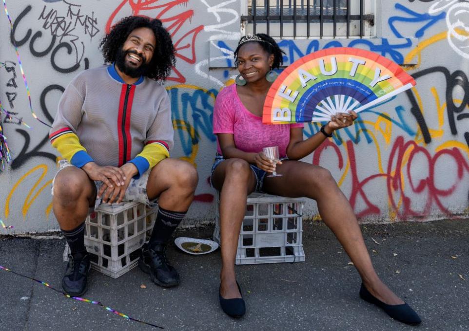 Photograph of a man and woman sitting on milk crates in front of a graffitied wall. The woman is holding a rainbow fan with the word 'Beauty.'