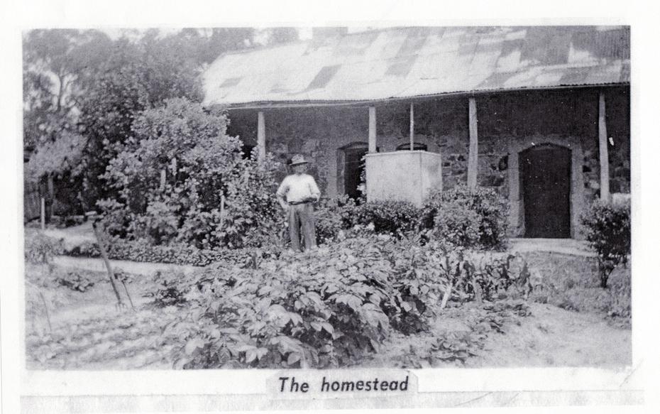 Black and white photo depicting a man standing in front of a stone cottage with vegetable garden