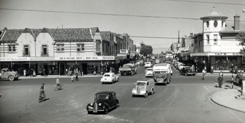 Black and white photograph showing cars driving past buildings on Whitehorse Road.