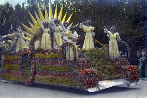 Hand coloured photograph of ladies waving on a float decorated with flowers and a sun.