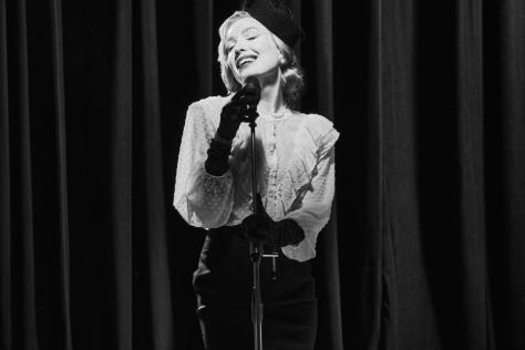 Black and white photo of woman dressed in hat and gloves singing into a microphone, stage curtain in background.