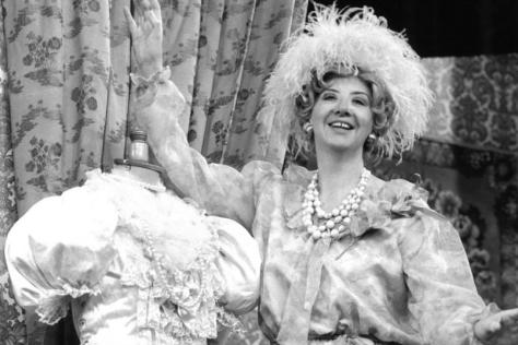 Black and white photo of actress on stage in a feather hat and pearls next to a costume mannequin