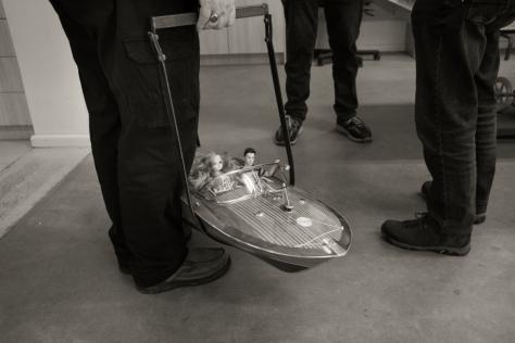 Black and white photo of a man holding a model speed boat with dolls driving 