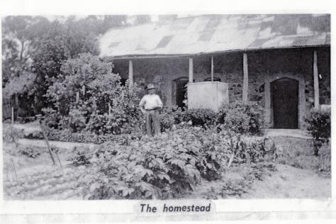 Black and white photo depicting a man standing in front of a stone cottage with vegetable garden