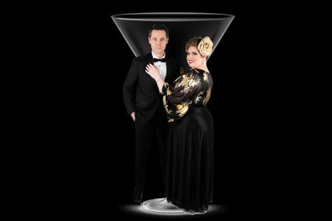 A man and a woman in black tie attire stand in front of a martini glass. 