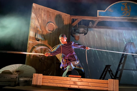 A masculine person in traditional Chinese clothing holds two ropes together in thick stage fog.