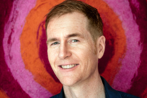 A man smiles at the camera, with pink and red circles behind him.
