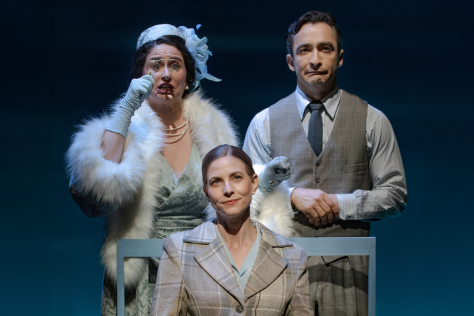 One woman sits on a chair while another woman stands behind, dressed in gloves and a hat, and a man in a tie and vest. 