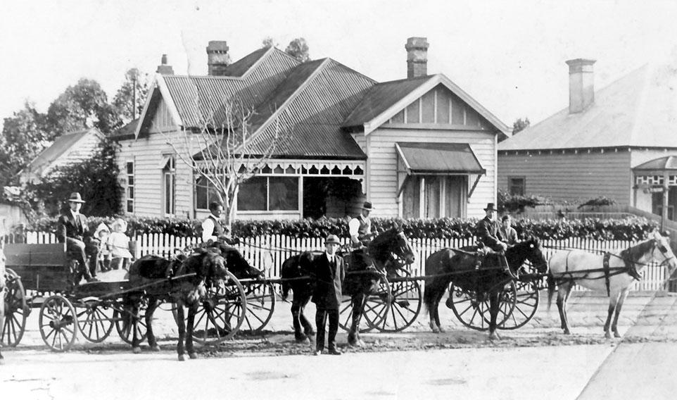 Black and white photograph of various horse and carts against suburban backdrop of a house